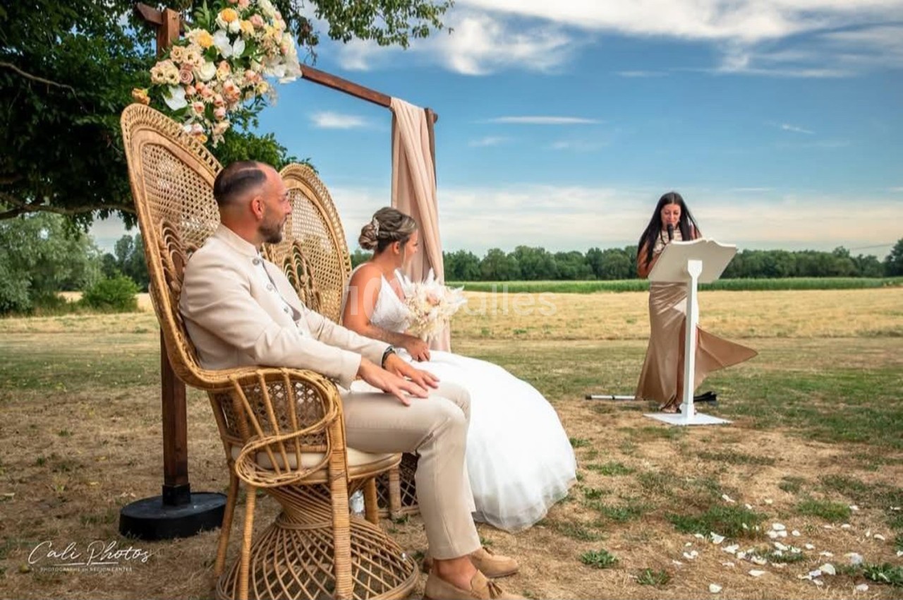 Un couple assis sur des fauteuils en osier écoute une femme prononçant un discours lors d'une cérémonie en plein air.