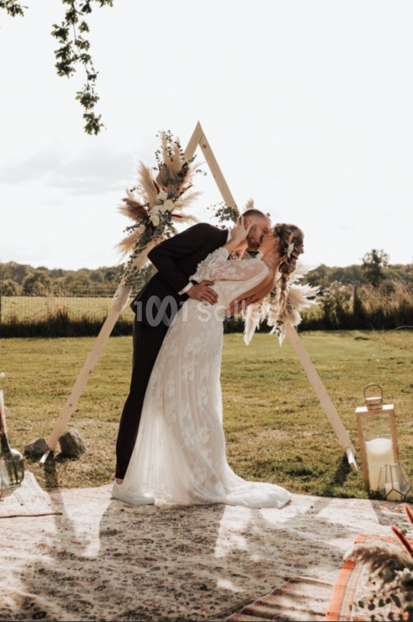 Un couple en tenue de mariage s'embrasse devant une arche décorée, dans un cadre champêtre en plein air.