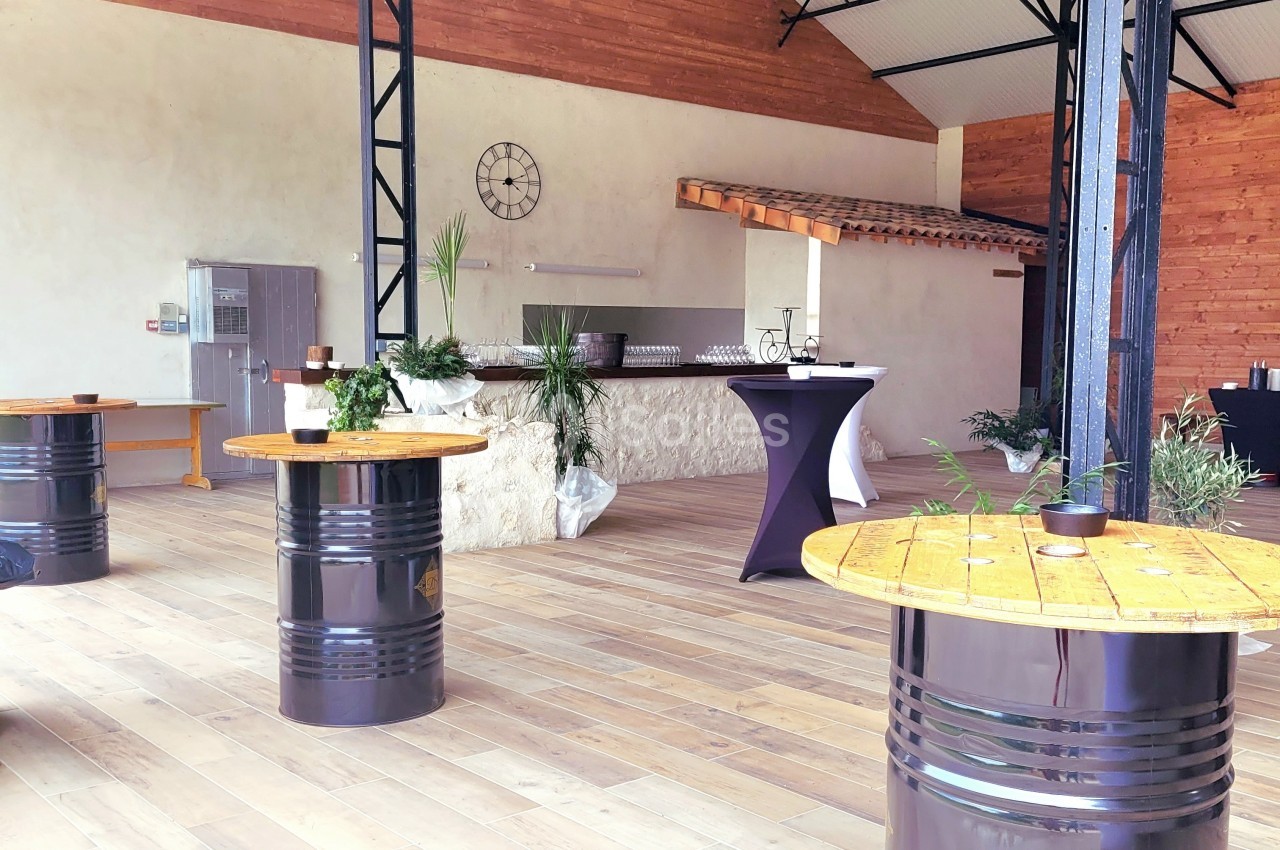 Salle lumineuse avec tables en tonneaux, bar en pierre, plafond haut et décorations végétales.