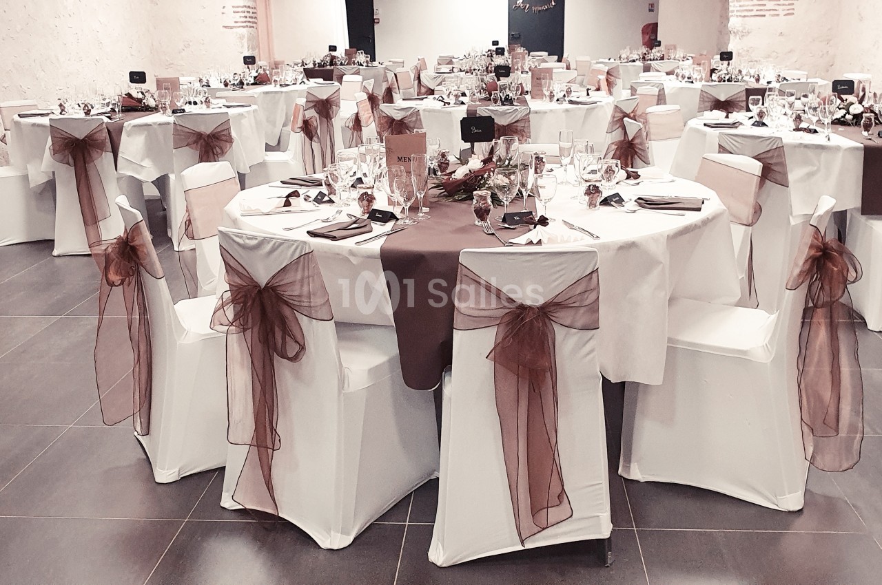 Salle de réception décorée avec des tables rondes, nappes blanches et nœuds en tissu marron sur les chaises.