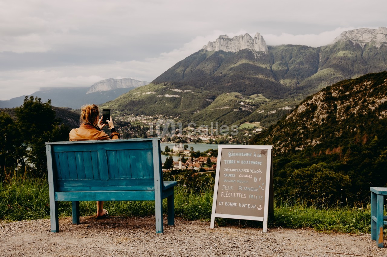 Une femme assise sur un banc bleu photographie un paysage montagneux avec une vallée et un panneau en bois à côté.