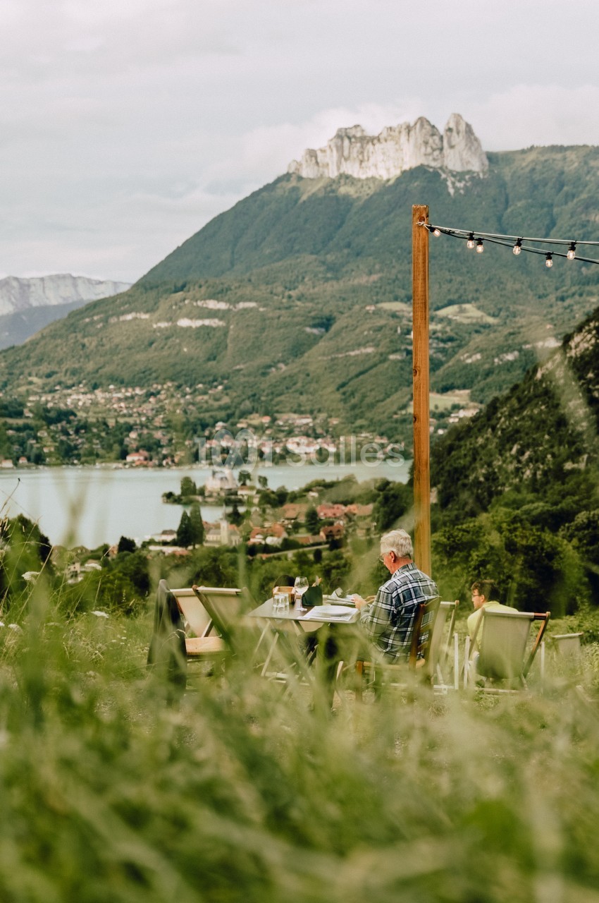 Un homme assis à une table en plein air, face à un paysage de montagne, d'un lac et d'une vallée verdoyante.