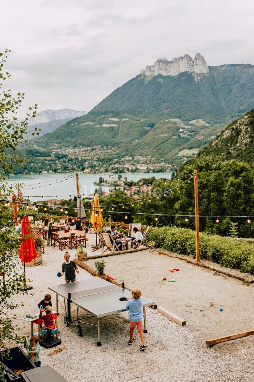 Enfants jouant au ping-pong sur une terrasse avec vue sur un lac, des montagnes et des tables de café en arrière-plan.
