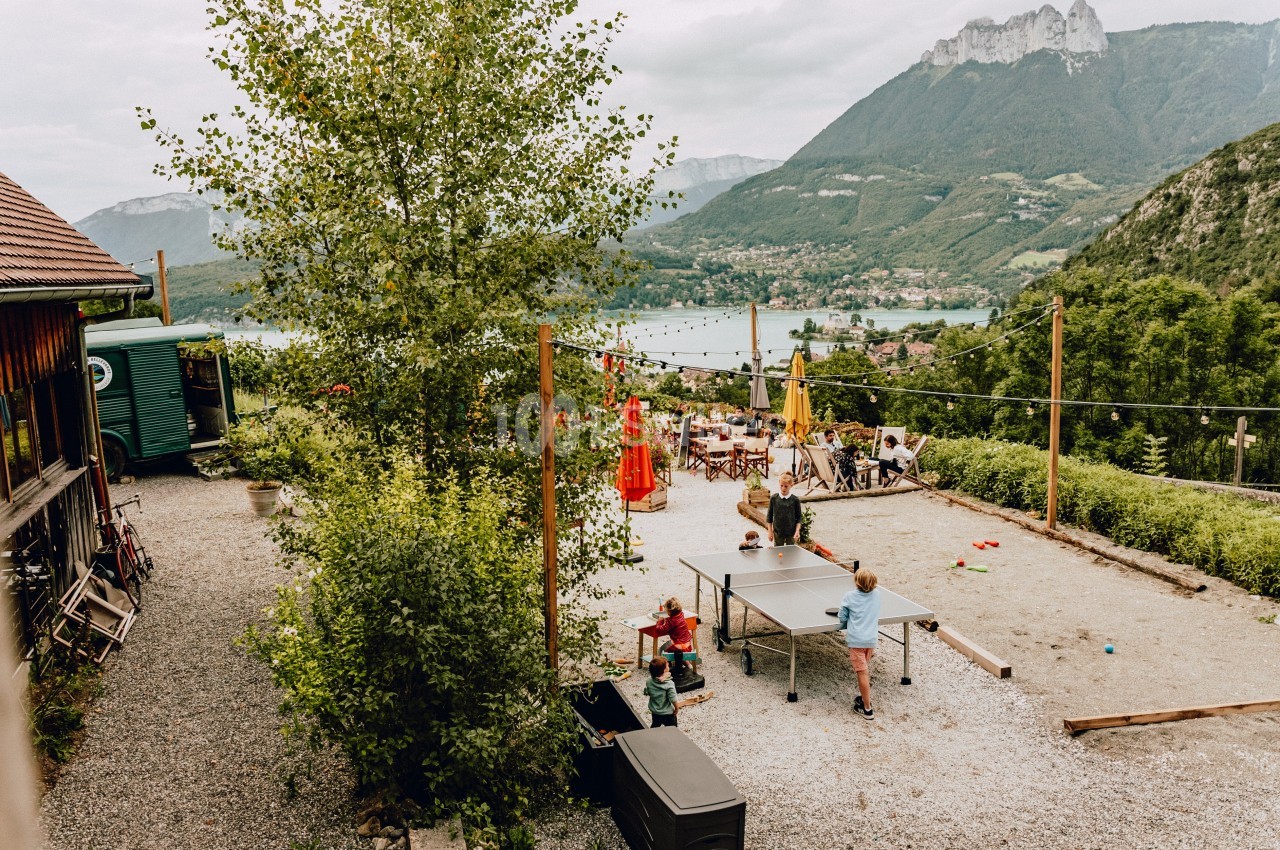 Enfants jouant au ping-pong dans une cour extérieure avec vue sur un lac et des montagnes verdoyantes en arrière-plan.