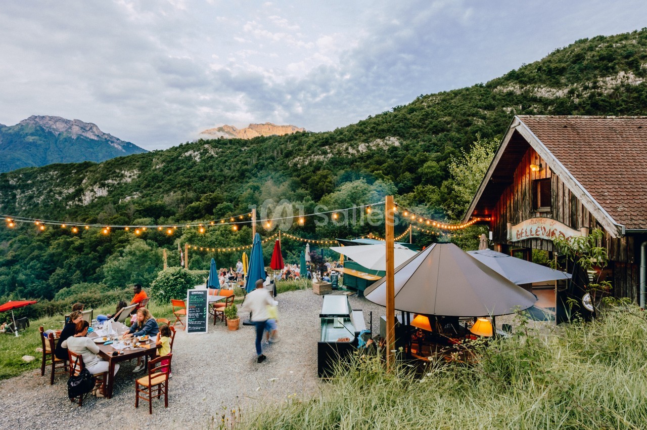 Terrasse extérieure d'un restaurant rural avec des tables, des parasols et des guirlandes lumineuses, entourée de montagnes.