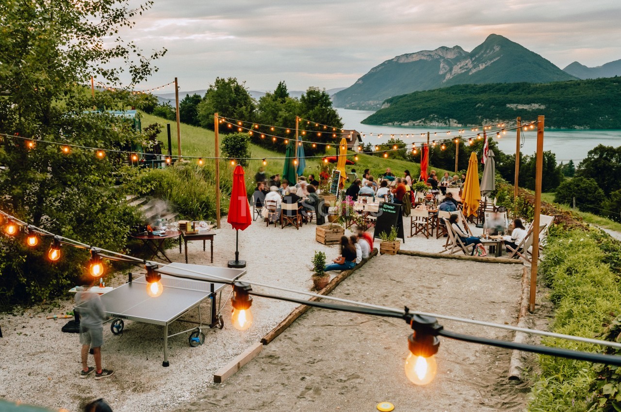 Terrasse extérieure animée avec tables, guirlandes lumineuses, vue sur un lac et montagnes en arrière-plan.