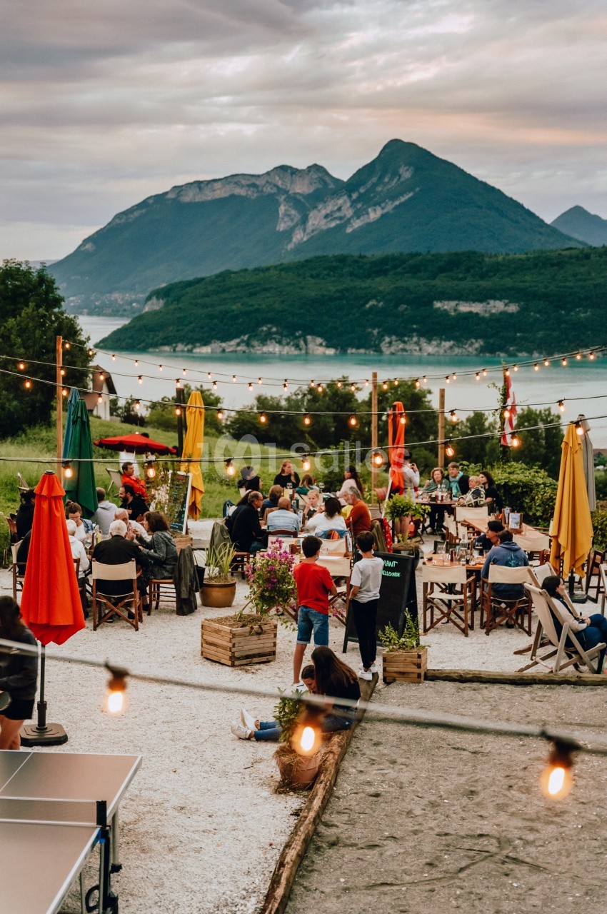 Terrasse animée en plein air avec des tables, des personnes et des lumières suspendues, vue sur un lac et des montagnes.