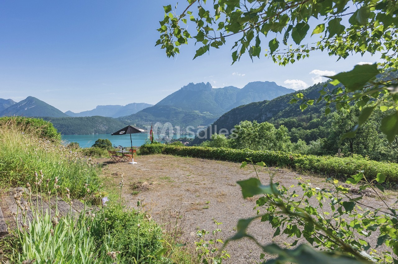 Vue sur un lac entouré de montagnes, avec un parasol noir sur une terrasse ensoleillée bordée de végétation.