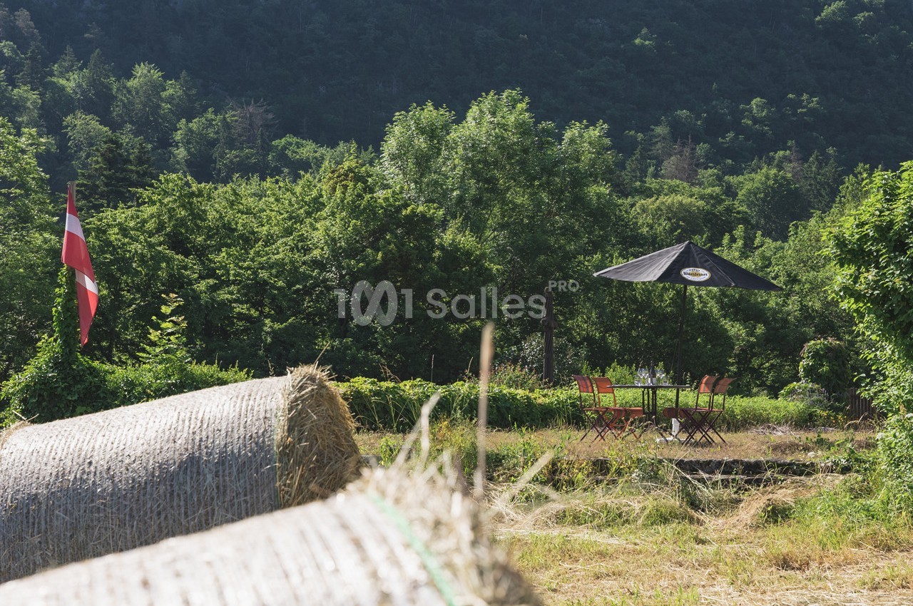 Champs avec bottes de foin, table et chaises sous un parasol noir, entourés de verdure et montagnes en arrière-plan.