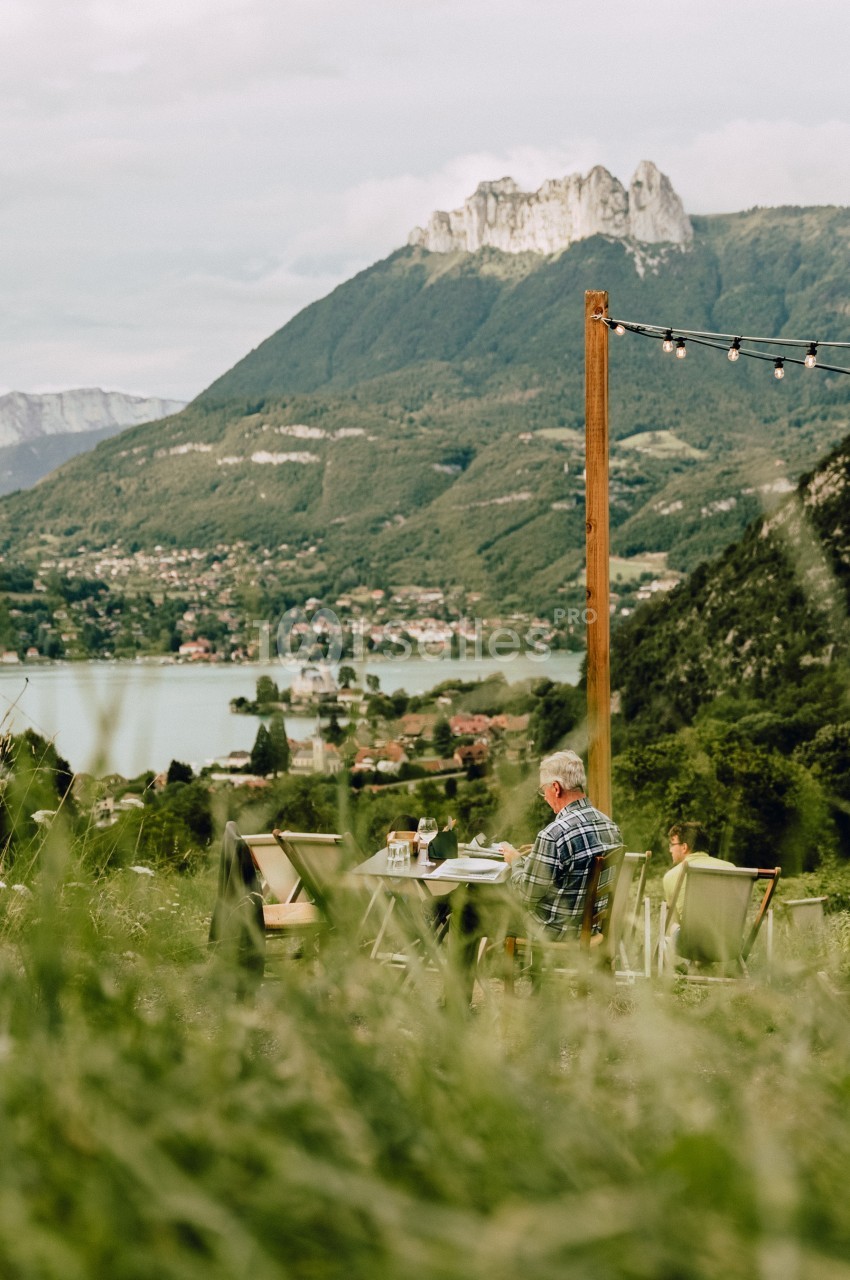Un homme assis à une table en plein air, face à un paysage de montagne, d'un lac et d'une vallée verdoyante.