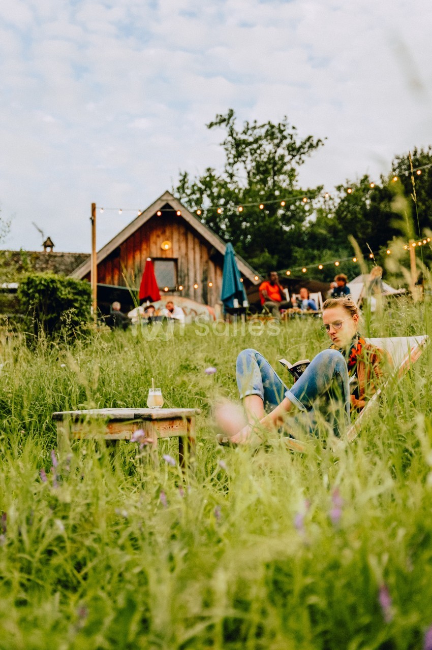 Une femme assise dans un champ verdoyant près d'une table basse, avec une maison en bois et des guirlandes lumineuses en…