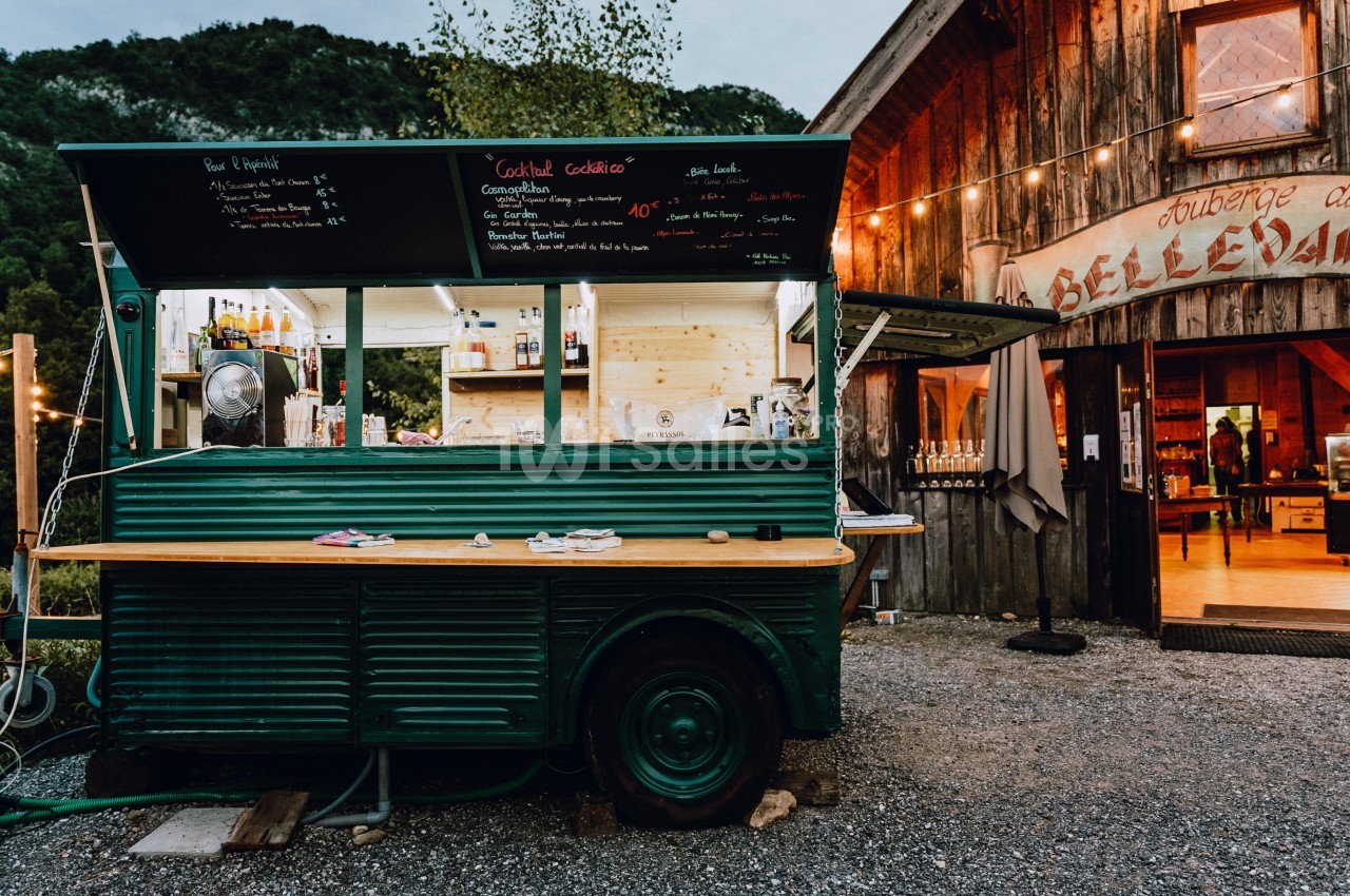 Food truck vert stationné devant une auberge en bois, avec menu écrit à la craie et ambiance extérieure rustique.