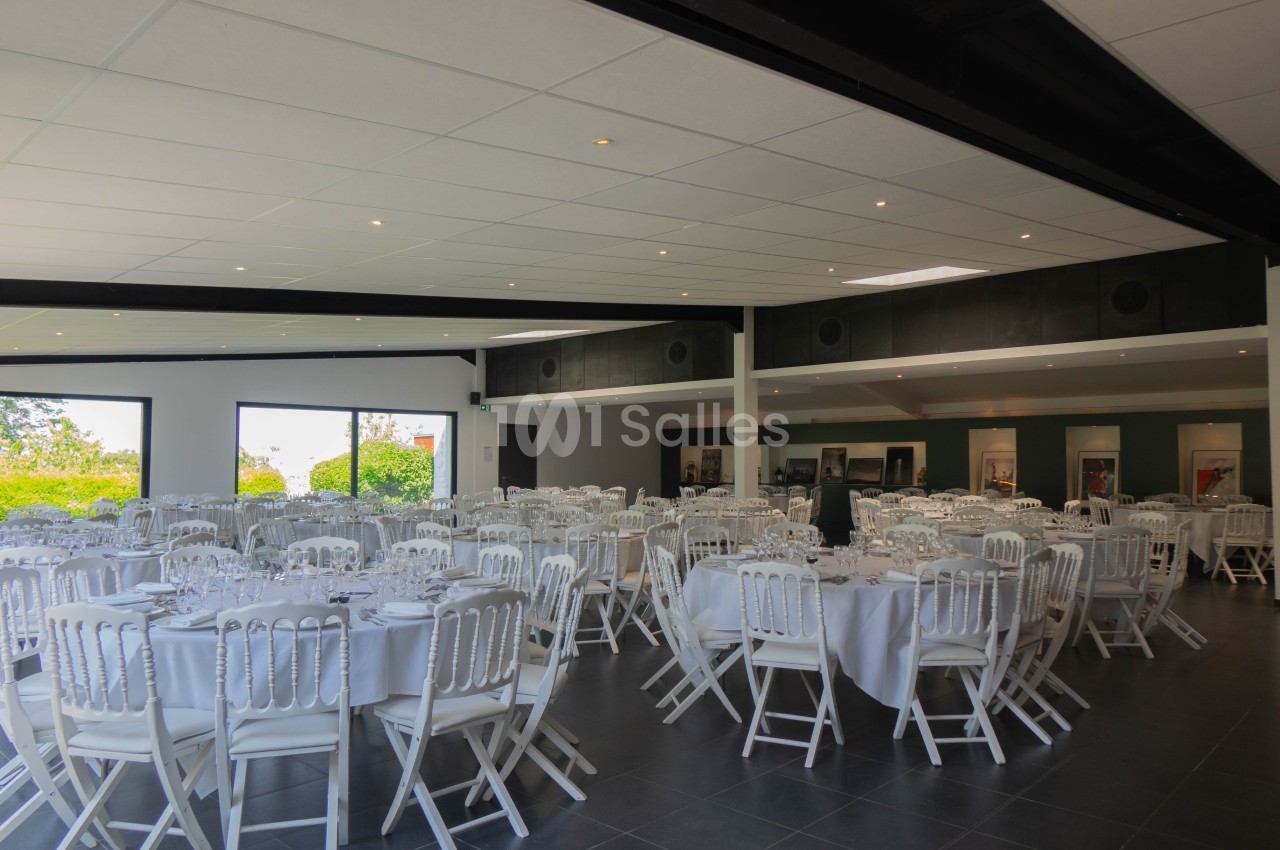 Salle de réception lumineuse avec des tables rondes dressées et des chaises blanches, vue sur l'extérieur verdoyant.