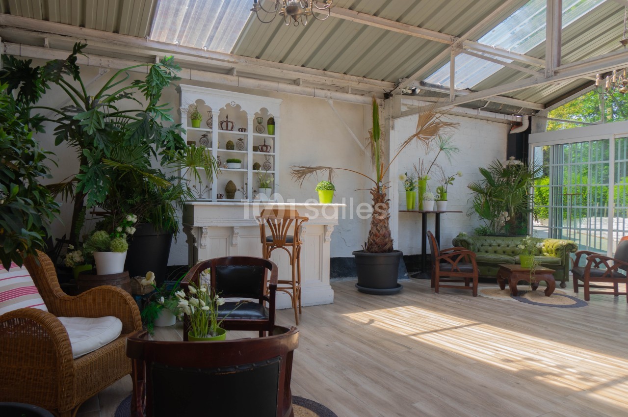 Salle lumineuse avec toit en verre, décorée de plantes, meubles en rotin et bois, et un bar blanc central.