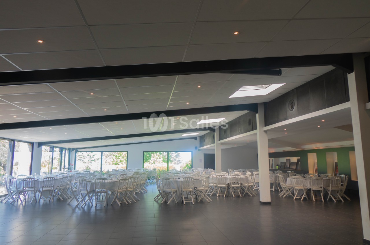 Salle spacieuse avec des tables rondes et des chaises blanches, éclairée par de grandes baies vitrées.
