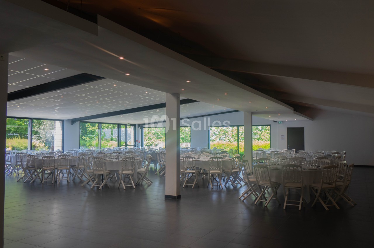 Salle de réception lumineuse avec tables rondes dressées et grandes baies vitrées donnant sur un jardin.