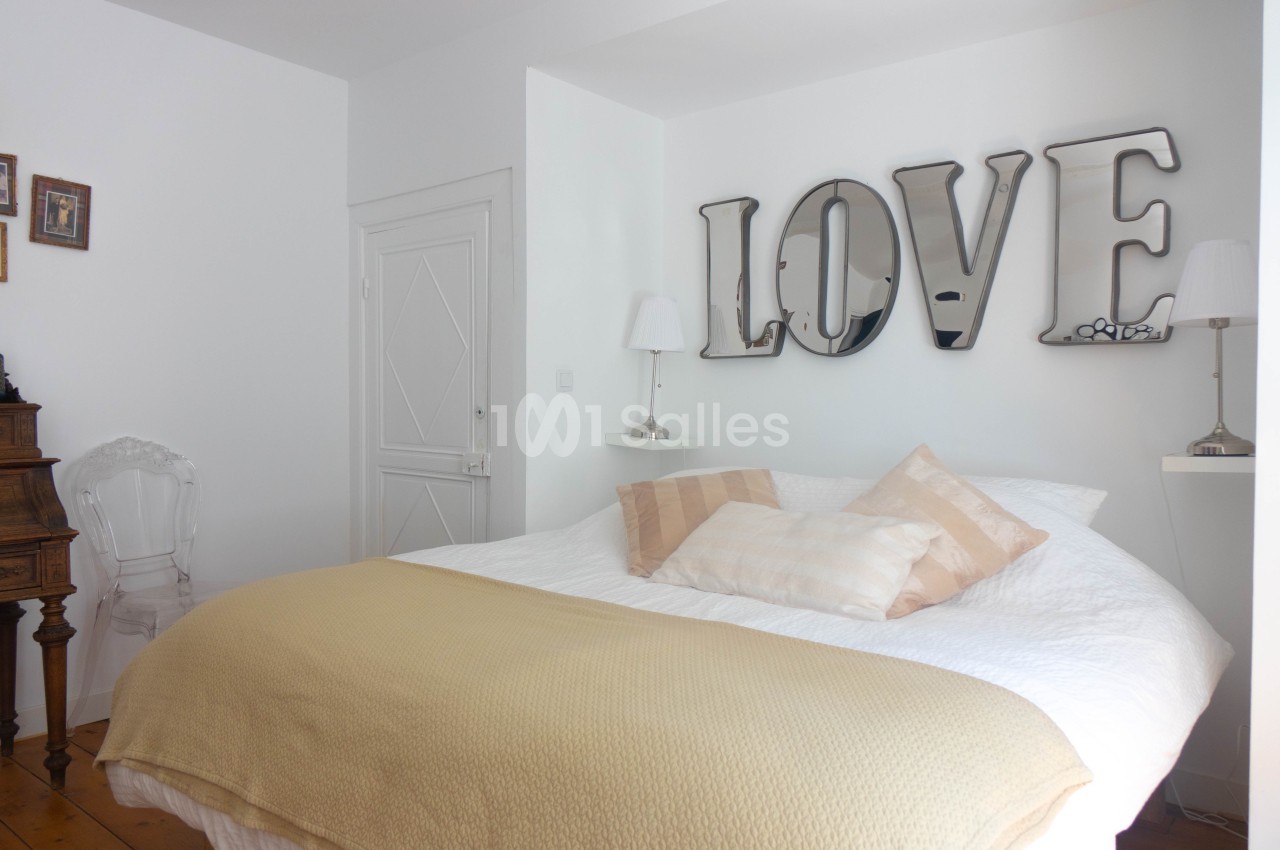 Lit double avec couverture beige, coussins assortis, mur blanc décoré de lettres métalliques formant ’LOVE’.