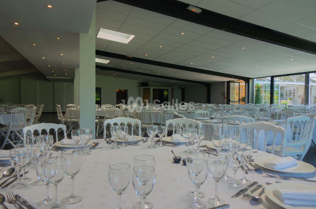 Salle de réception lumineuse avec tables dressées, nappes blanches, verres et couverts, prête pour un événement.