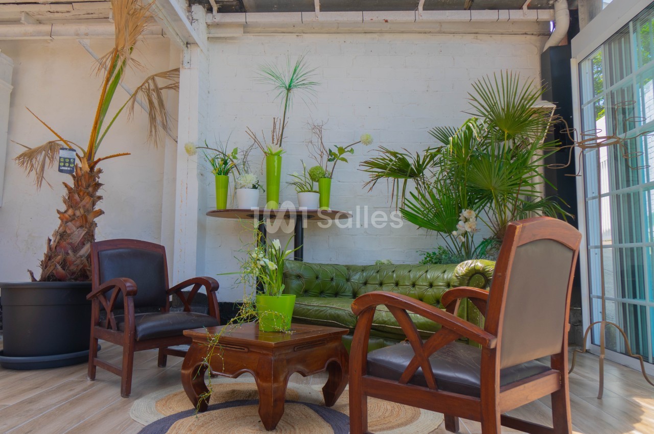 Salon lumineux avec canapé vert, fauteuils en bois, table basse et plantes décoratives contre un mur blanc.