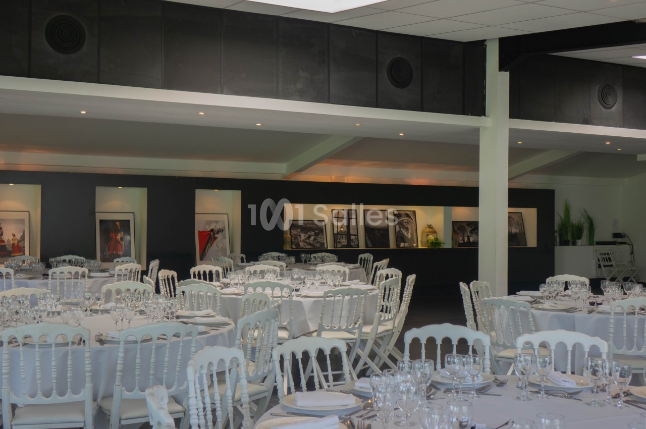 Salle de réception avec tables dressées, chaises blanches et décorations murales comprenant des photographies encadrées.