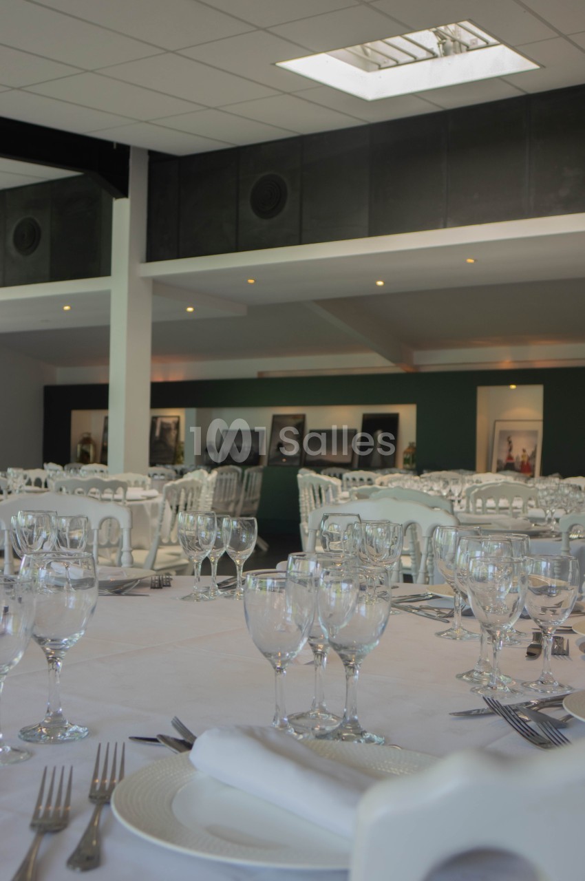 Salle de réception lumineuse avec tables dressées, nappes blanches, verres et couverts, décor sobre en arrière-plan.