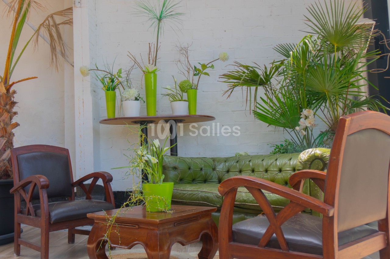 Salon lumineux avec canapé vert, fauteuils en bois, table basse et plantes vertes décoratives contre un mur blanc.