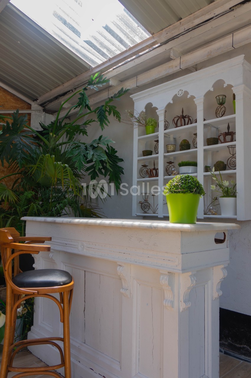 Comptoir blanc avec tabouret en bois, étagère décorative en arrière-plan et plantes vertes sous un toit partiellement ouvert.
