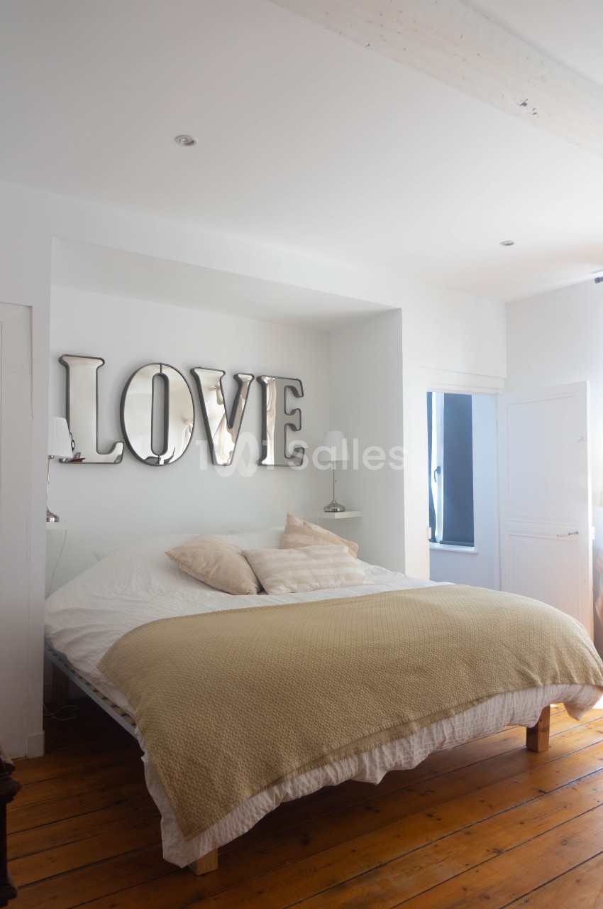 Chambre lumineuse avec un lit double, coussins beiges, parquet en bois et décoration murale ’LOVE’ en lettres métalliques.