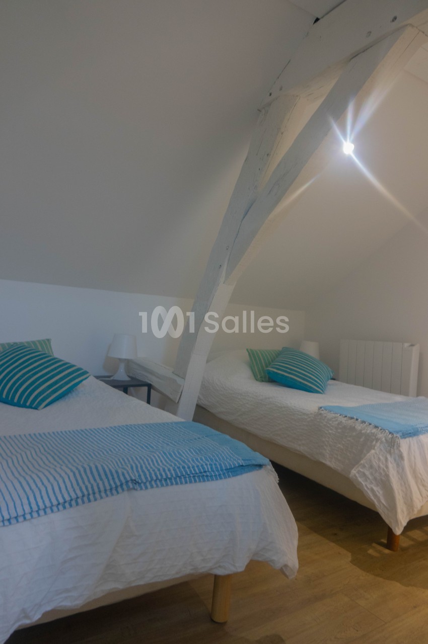 Deux lits simples avec draps blancs et coussins rayés bleus dans une chambre mansardée lumineuse.