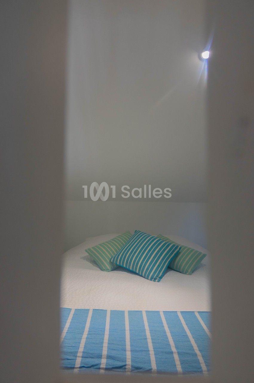 Lit avec une couverture bleue rayée et des coussins assortis, vu à travers une ouverture dans un espace lumineux.