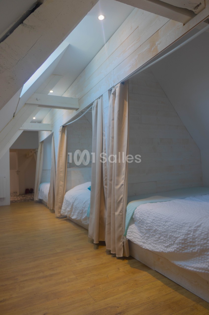 Chambre mansardée avec lits simples alignés, séparés par des rideaux, et éclairée par des fenêtres de toit.