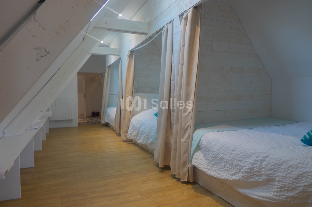 Chambre mansardée avec lits alignés séparés par des rideaux, parquet au sol et murs en bois clair.