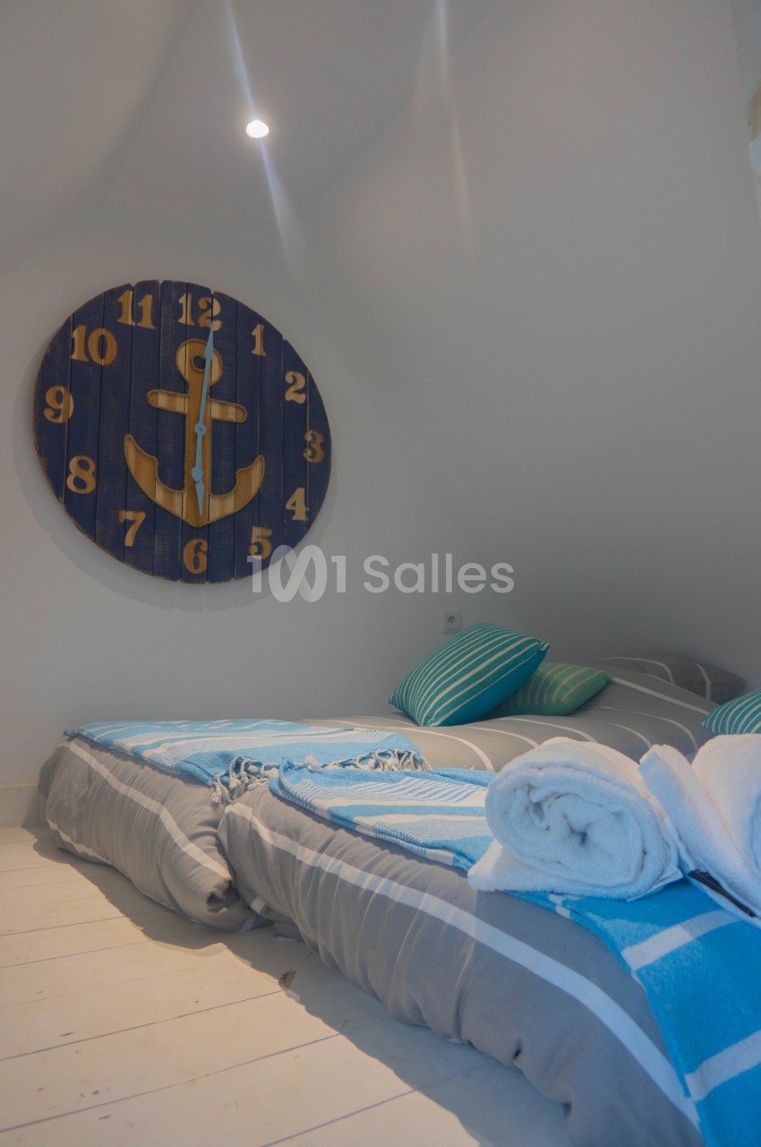 Deux lits simples avec draps bleus et gris, serviettes roulées, et une horloge murale en bois décorée d'une ancre.