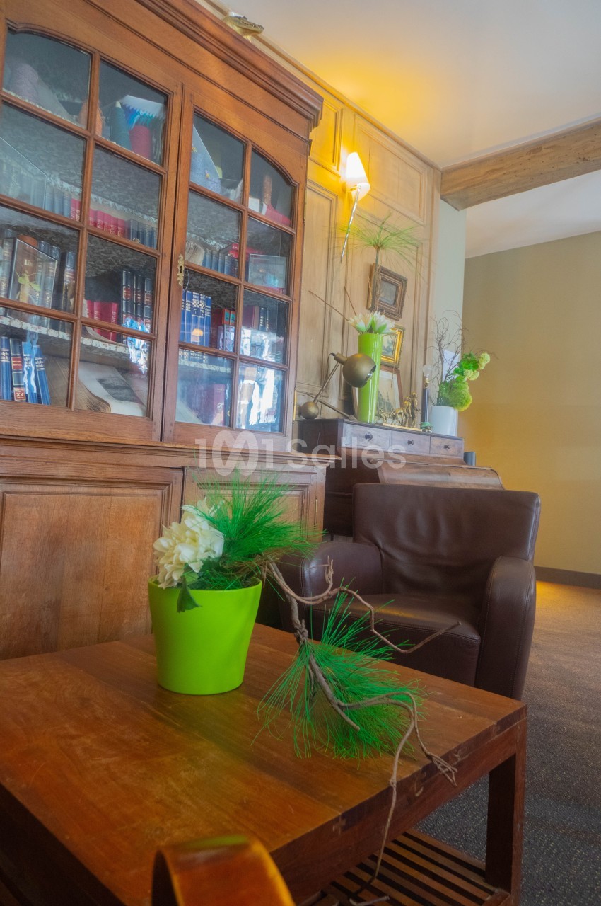 Salon avec fauteuil en cuir, table en bois, plante verte en pot et bibliothèque vitrée contenant des livres.