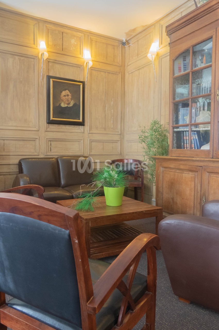 Salon avec fauteuils en cuir, table basse en bois, plante verte, bibliothèque et tableau sur un mur lambrissé.
