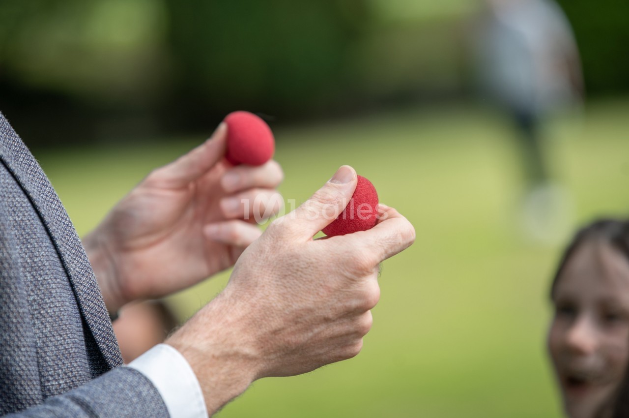 Deux mains tenant des balles rouges en mousse dans un jardin, avec des personnes floues en arrière-plan.