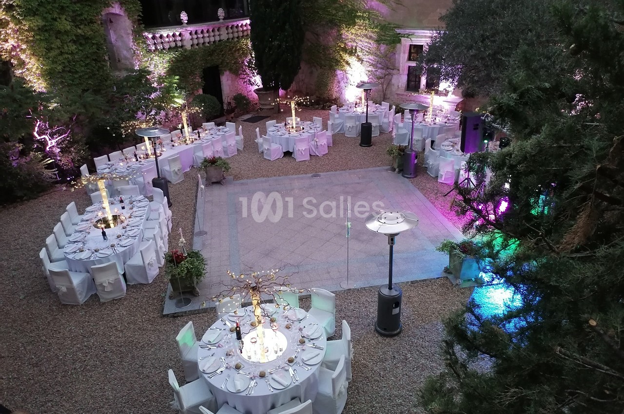 Cour intérieure aménagée pour un événement, avec tables rondes décorées, éclairages colorés et végétation environnante.