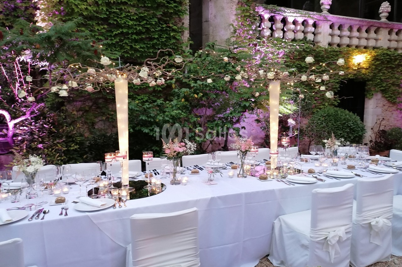 Table de réception décorée avec des fleurs, des bougies et des nappes blanches, dans un jardin avec murs recouverts de…