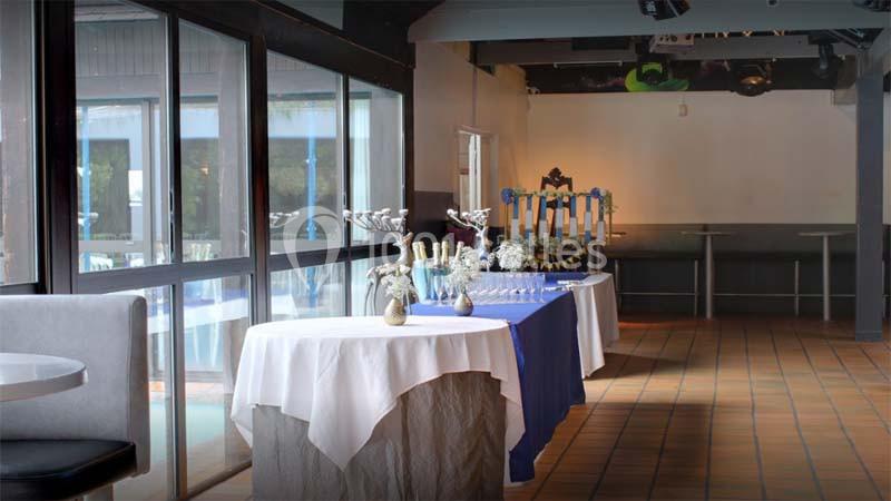 Salle de réception avec tables dressées, nappes blanches et bleues, décorations et verres alignés près de grandes baies…