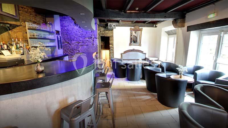 Intérieur d'un bar avec comptoir courbé, tabourets hauts, fauteuils noirs et murs en pierre apparente.