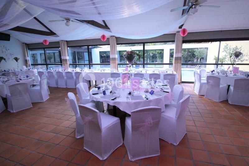 Salle décorée pour un événement avec tables rondes dressées, chaises blanches et décorations roses sous un plafond voilé.