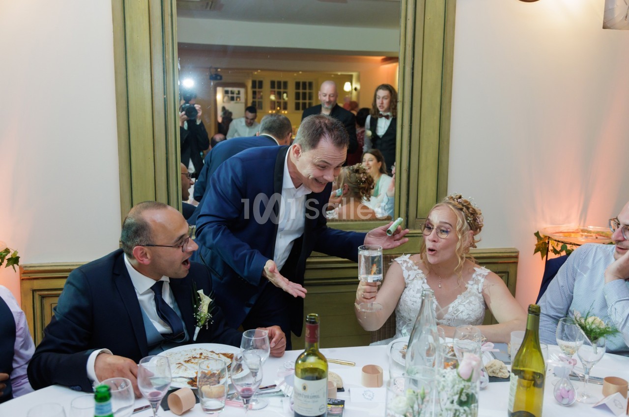 Un homme en costume plaisante avec une mariée tenant un verre, entourés d'invités à table lors d'une réception.