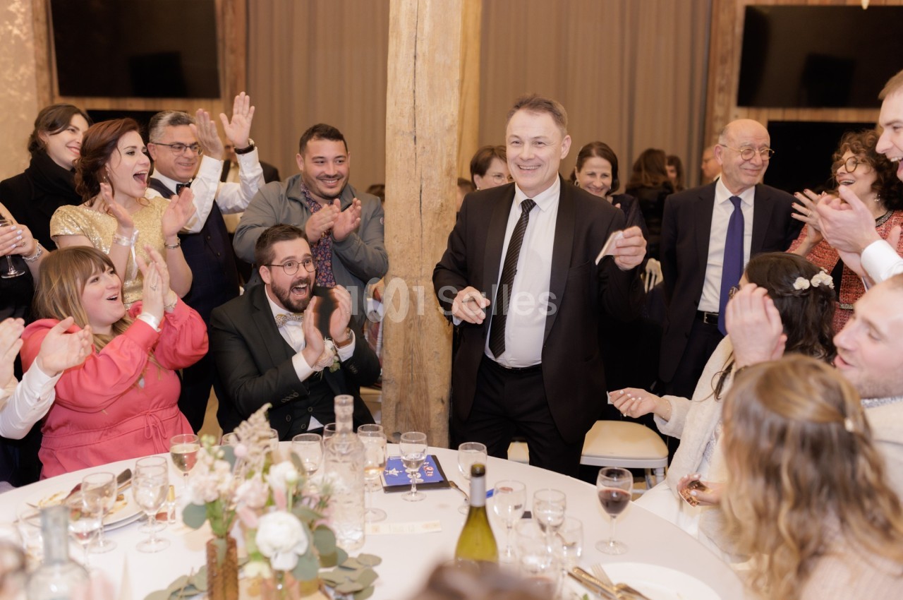 Un homme en costume debout au centre d'une salle, entouré de convives applaudissant autour d'une table décorée.
