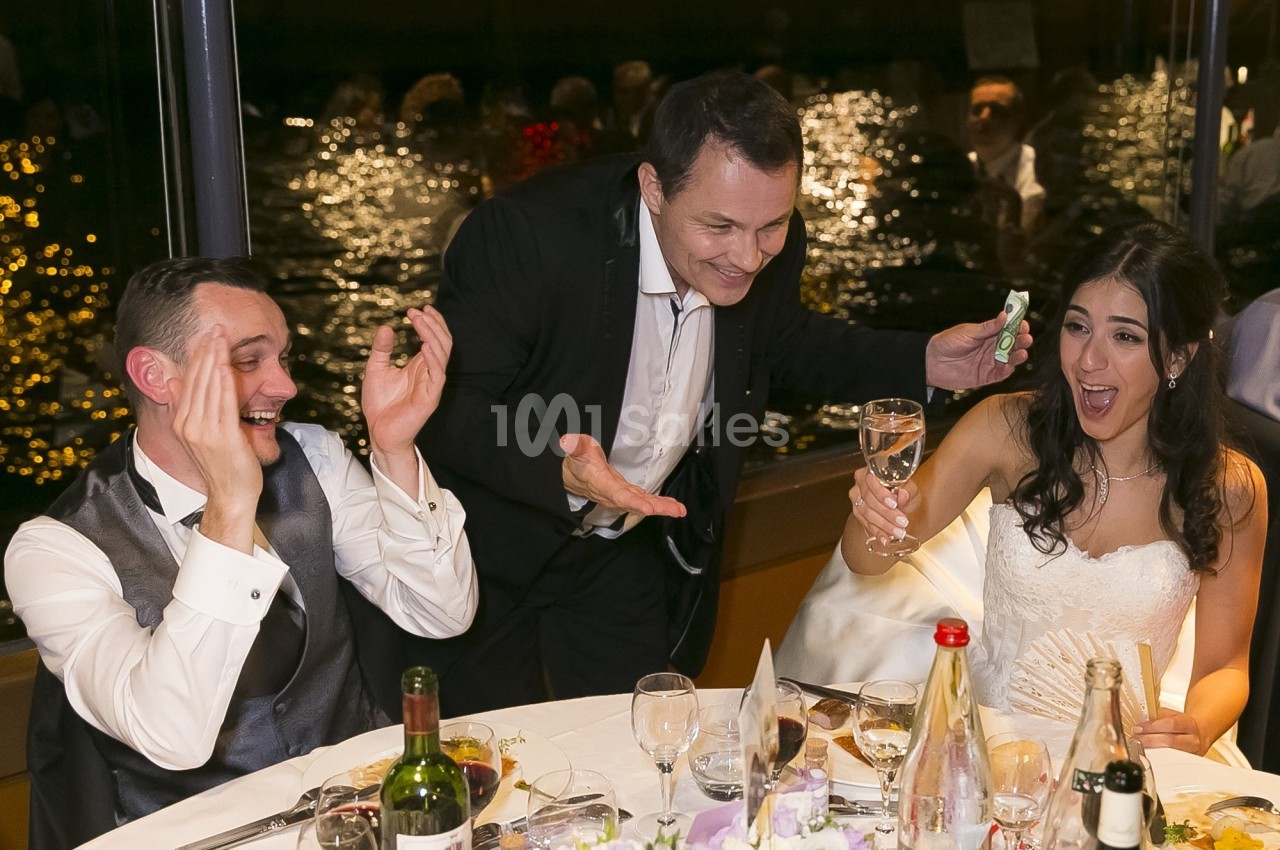 Un couple de mariés assis rit avec un invité debout qui tient un verre et un billet, lors d'une réception festive.