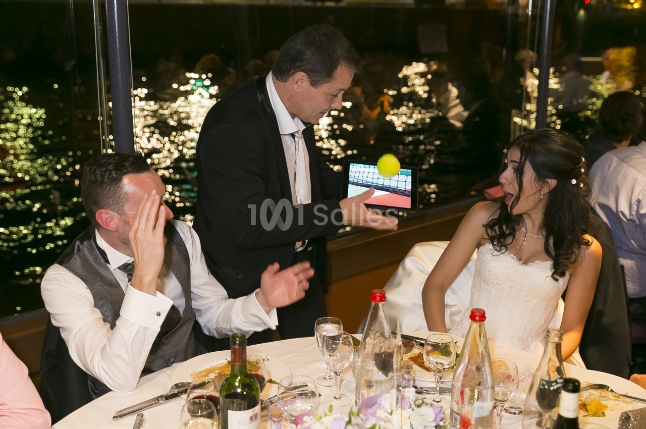 Un magicien montre un tour avec une balle et une tablette à des convives assis à une table lors d'un dîner.