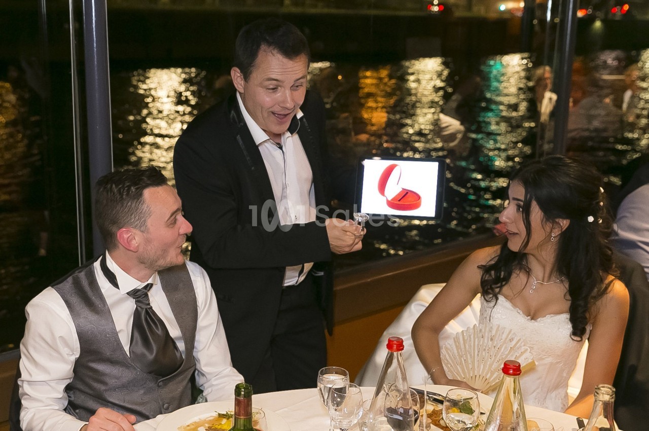Un magicien présente un tour avec une tablette à un couple assis à table lors d'un dîner près d'une rivière.
