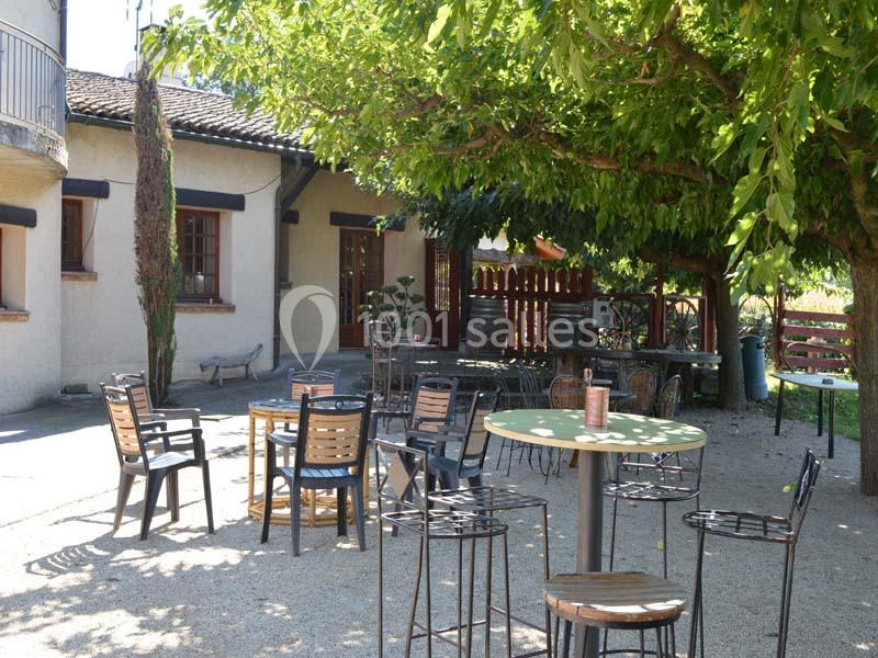 Terrasse ombragée avec tables et chaises en métal et bois, entourée d'arbres et d'une maison en arrière-plan.
