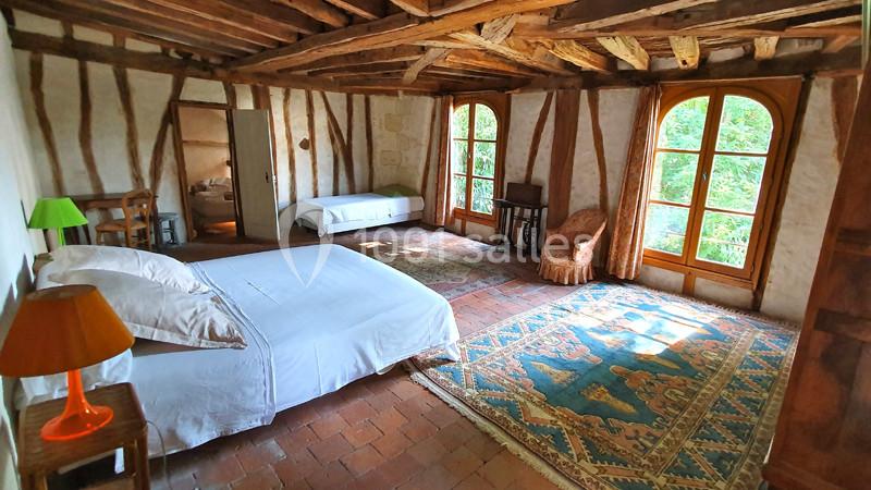 Chambre spacieuse avec poutres apparentes, lit double, lit simple, tapis coloré et grandes fenêtres donnant sur la verdure.