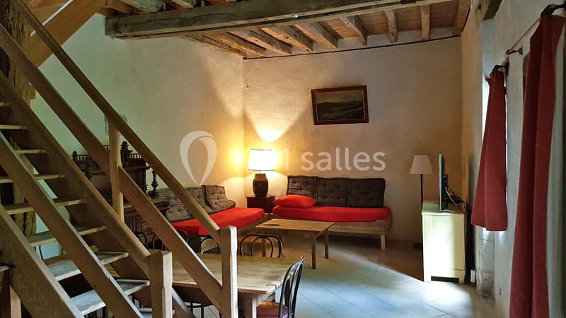 Salon rustique avec poutres apparentes, canapé rouge, lampes allumées, table en bois et escalier en bois.