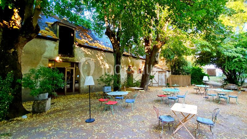 Cour extérieure ombragée avec tables et chaises en métal, entourée d'arbres et d'un bâtiment ancien en pierre.